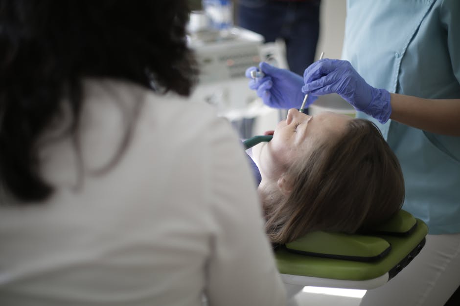 Crop anonymous female dentist in blue uniform and latex gloves using drill and mouth mirror while treating teeth of female patient lying in dental chair with opened mouth and closed eyes