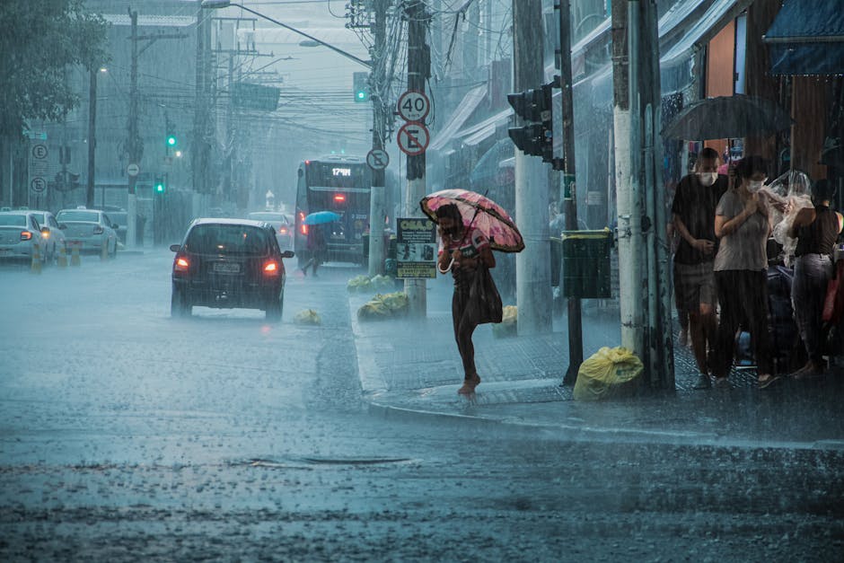 A busy urban street during heavy rain, with cars, pedestrians, and umbrellas.