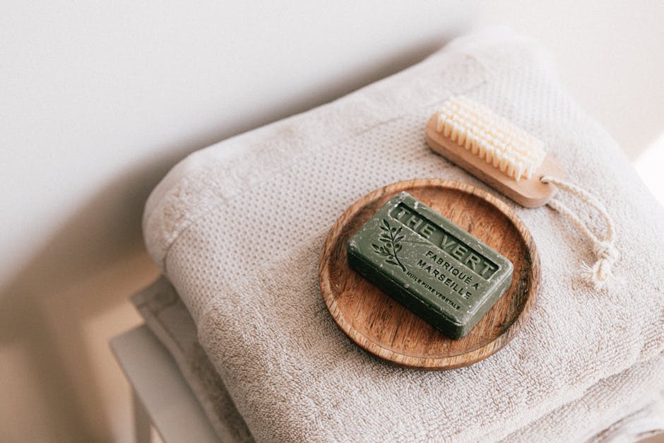From above composition of organic green natural soap on wooden holder and eco friendly brush placed on gray clean folded towels on white stool