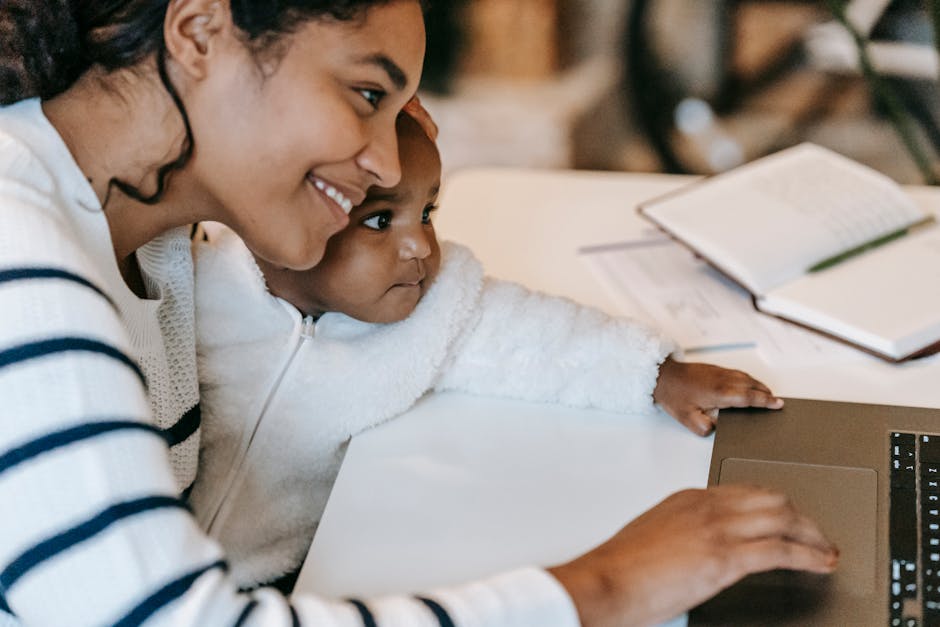 Side view of crop cheerful young black mother with curious infant in warm casual clothes using laptop sitting at table at home