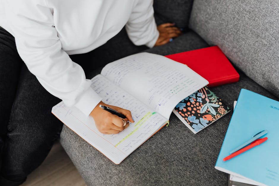 A woman in a white sweater writes notes on a notebook while studying on a sofa.
