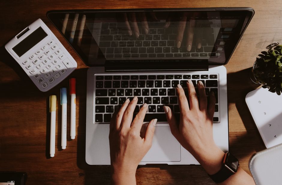 Hands typing on a laptop with a calculator and pens nearby on a wooden table.