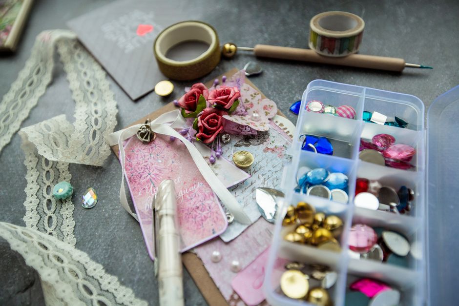 From above box of various colorful fancy stones with lace and adhesive tapes placed on table near handmade greeting card decorated with artificial flowers