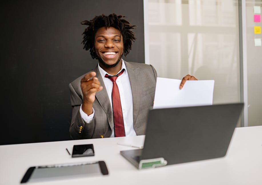 Smiling businessman holding paper at desk with laptop, embodying confidence and professionalism.