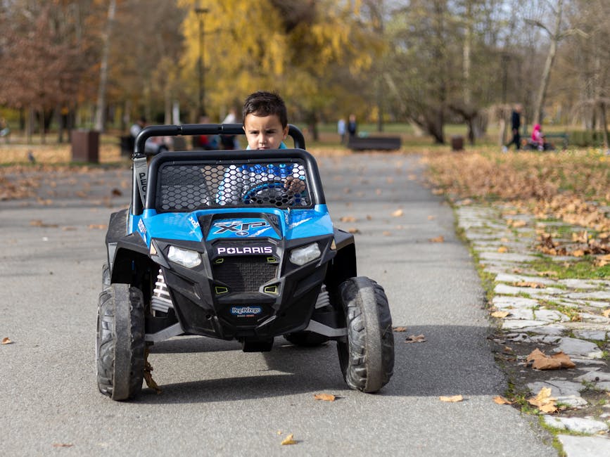 Young boy riding a blue electric toy car in a park on a sunny autumn day.