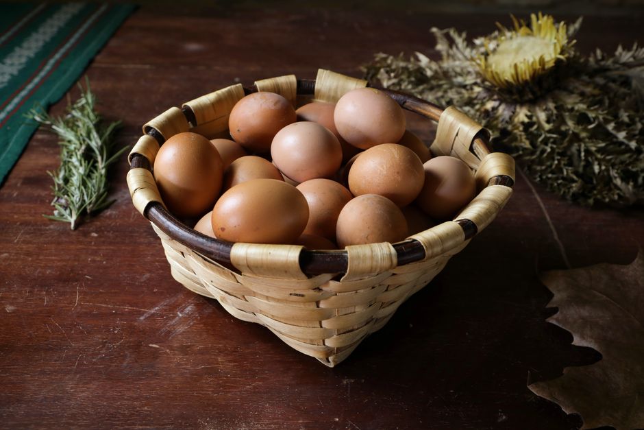 A rustic wicker basket filled with brown eggs on a wooden table, surrounded by herbs and dried flowers.