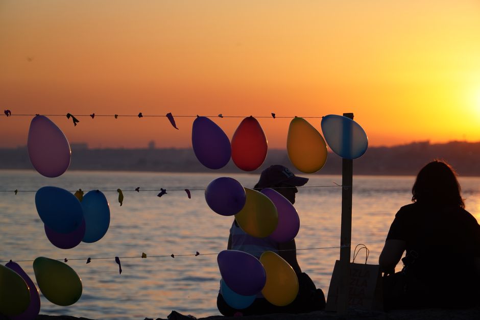 Two people sitting by colorful balloons on a beach during sunset.