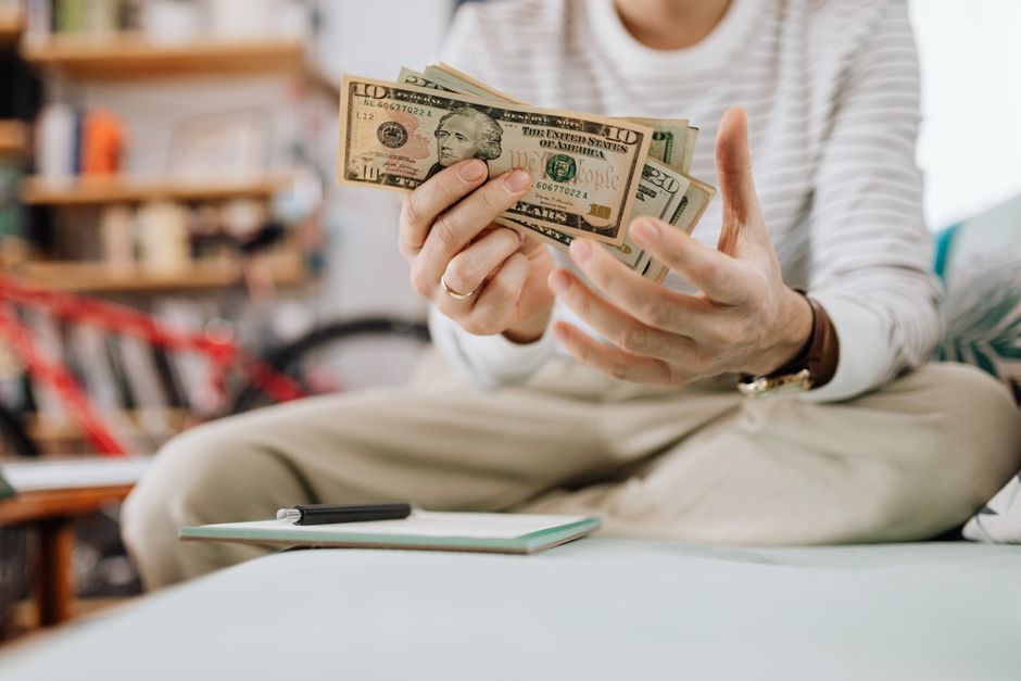 Man wearing casual clothes counts dollar bills while sitting on a sofa indoors.