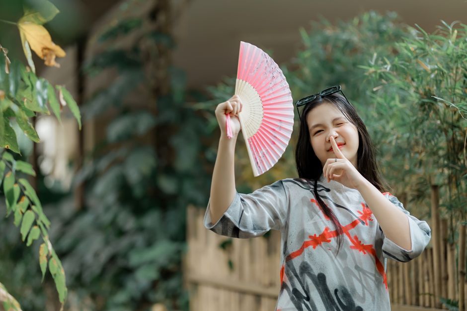 Smiling woman holding a pink fan in a lush garden setting, showcasing a playful mood.