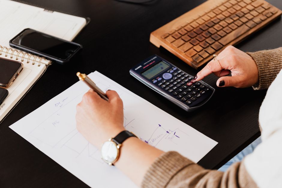 From above crop unrecognizable female accountant wearing stylish watches using calculator and writing down information on paper while working at desk with gadgets