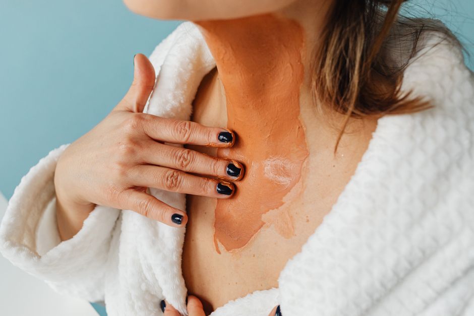 Close-up of woman applying clay mask on neck in spa robe for skincare routine.