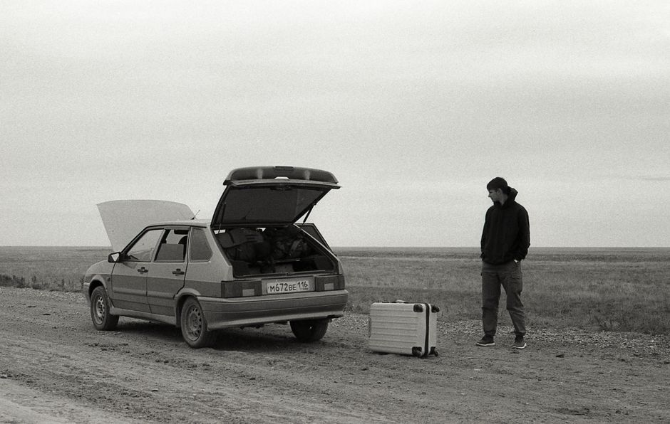 A man stands by a broken down car on a dirt road with luggage in a desolate, rural setting.