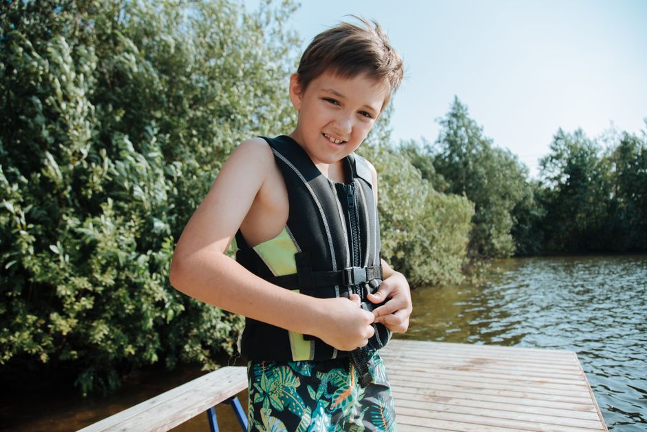 Happy child in a life jacket smiling at the camera while standing on a lakeside jetty.