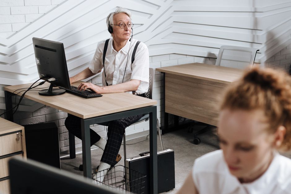Senior call center agent using a headset and computer in a modern office setting.