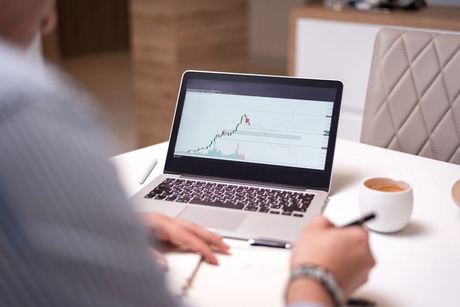 A person in a home office analyzing stock market data on a laptop with a coffee cup beside them.