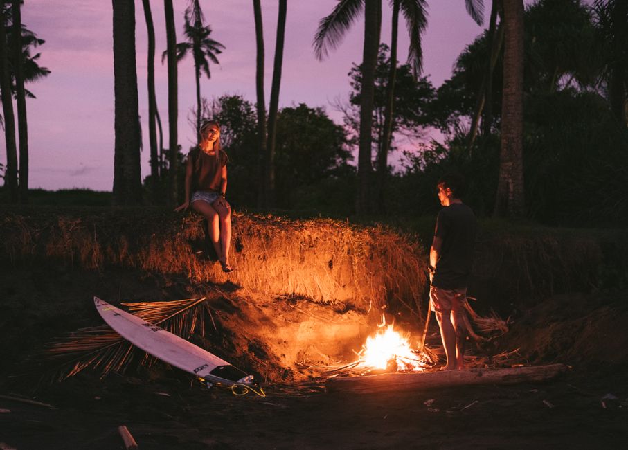 Full body of young unrecognizable man and woman in summer outfits chilling together near bonfire during romantic picnic on beach against amazing purple sunset sky
