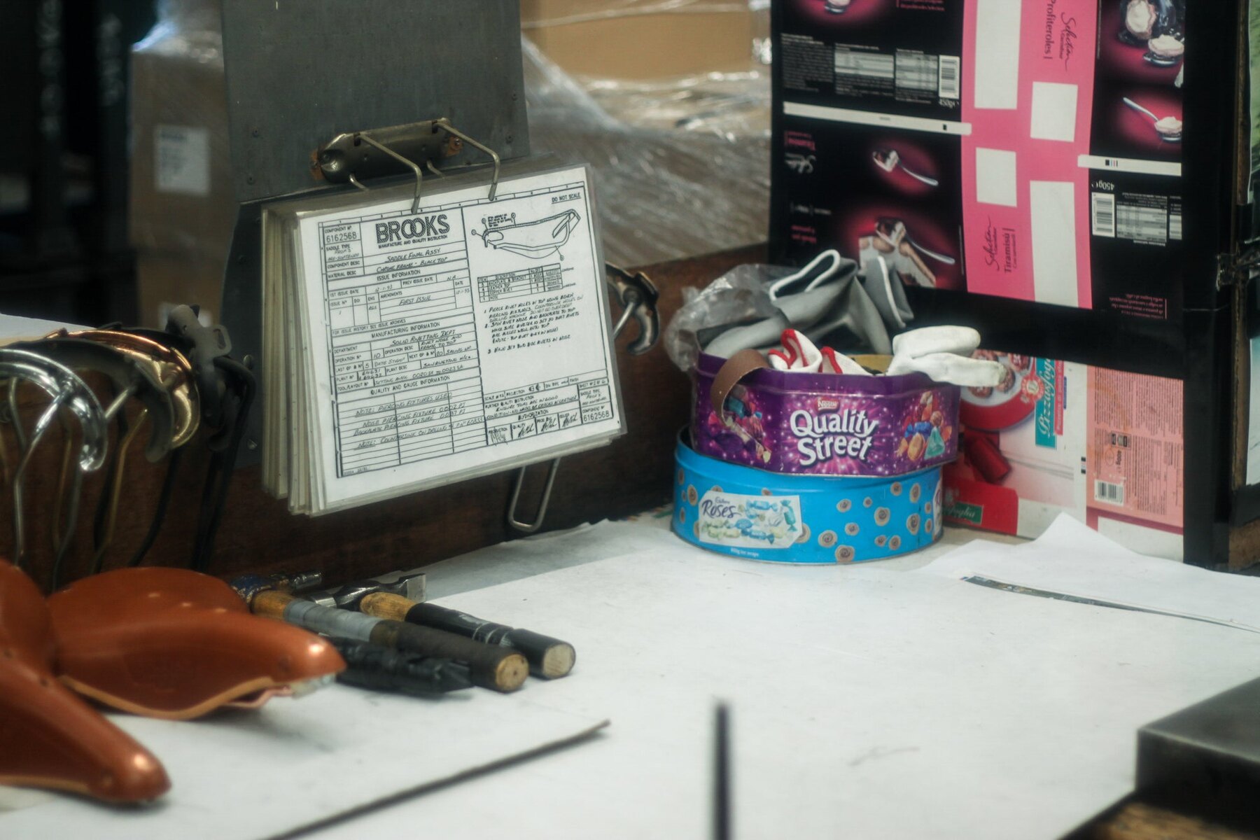 A working bench at Smethwick: leather tops stacked at the left, hand punches and drifts laid out, a job card on a clipboard, and loose components kept in biscuit tins. The leather saddles are still made here; the Cambium is made in Italy.