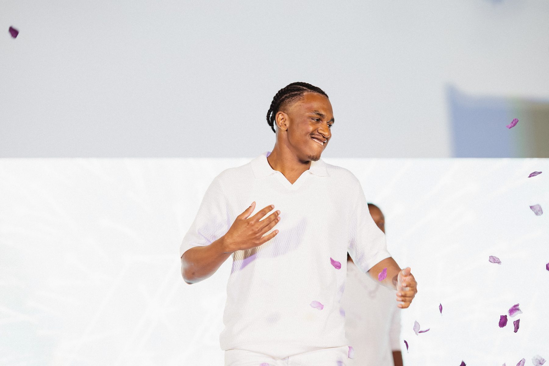 A young man smiles on stage at TheGifted Arts as purple petals fall around him.