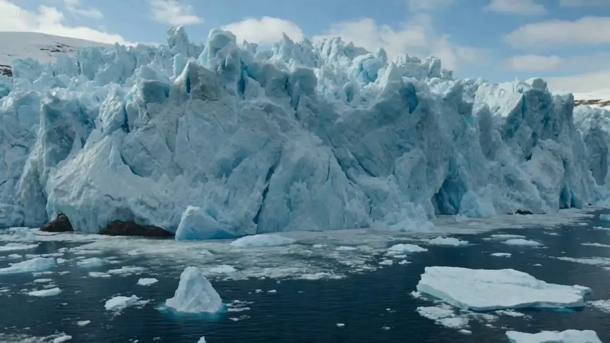 A giant glacier wall collapses into a fjord beside a coas...