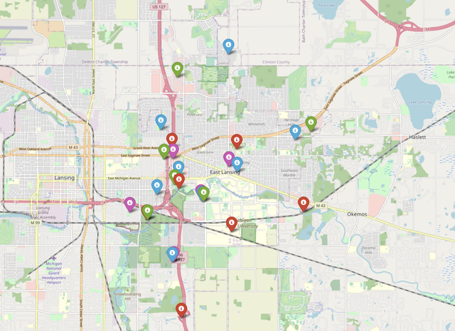 A street map of the Lansing and East Lansing area with cell-tower locations shown as colored map pins (green, red, blue, and purple), where the colors represent the different frequencies assigned by the graph-coloring algorithm.