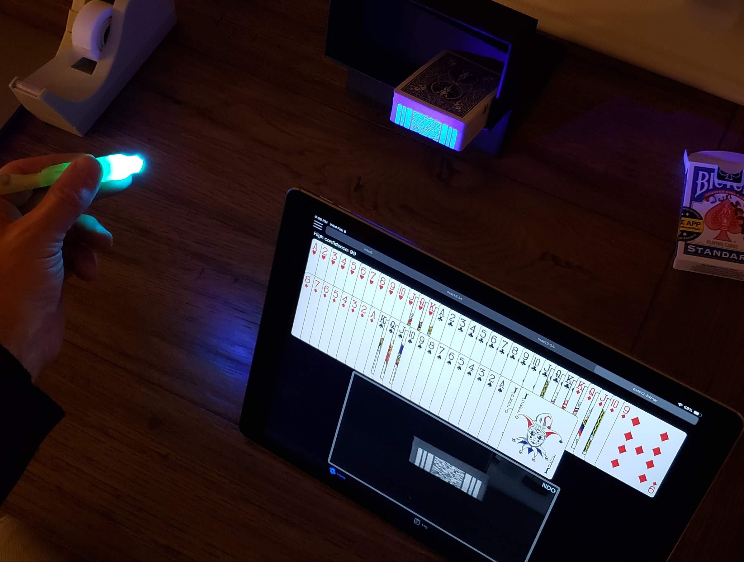 A deck of cards with marks on the edges of cards that are glowing brightly under the light of a UV pen light. Next to the deck is an iPad showing the deck from it's camera's perspective.