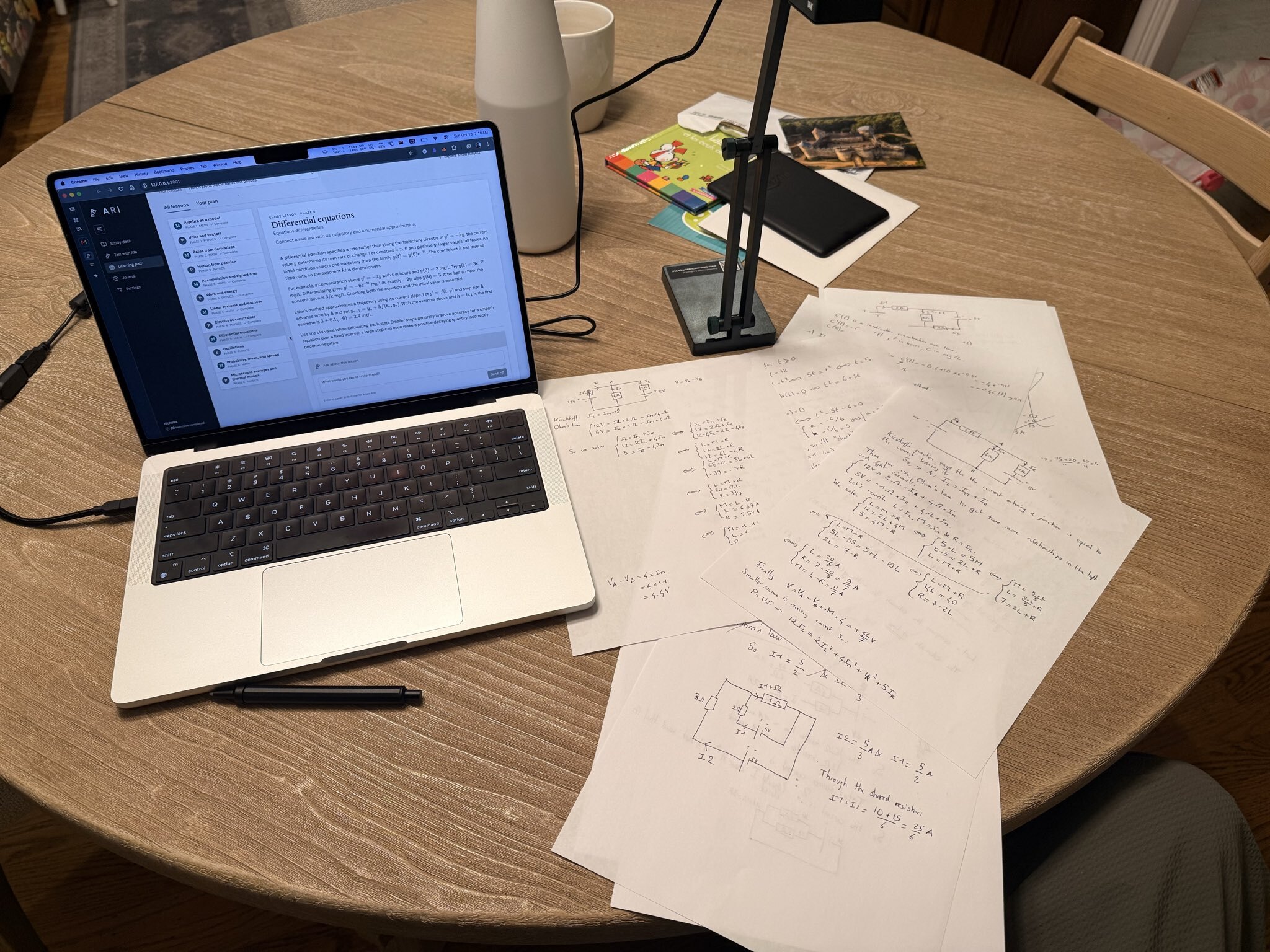 Laptop running Ari beside a document camera and a page of handwritten maths and physics work