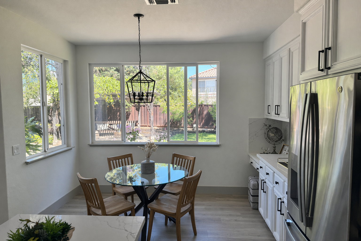 The same nook regenerated with four wooden slat-back chairs: the lantern pendant is larger and hangs higher, the garden beyond the window has been redrawn, and the frame is wider than the original photograph