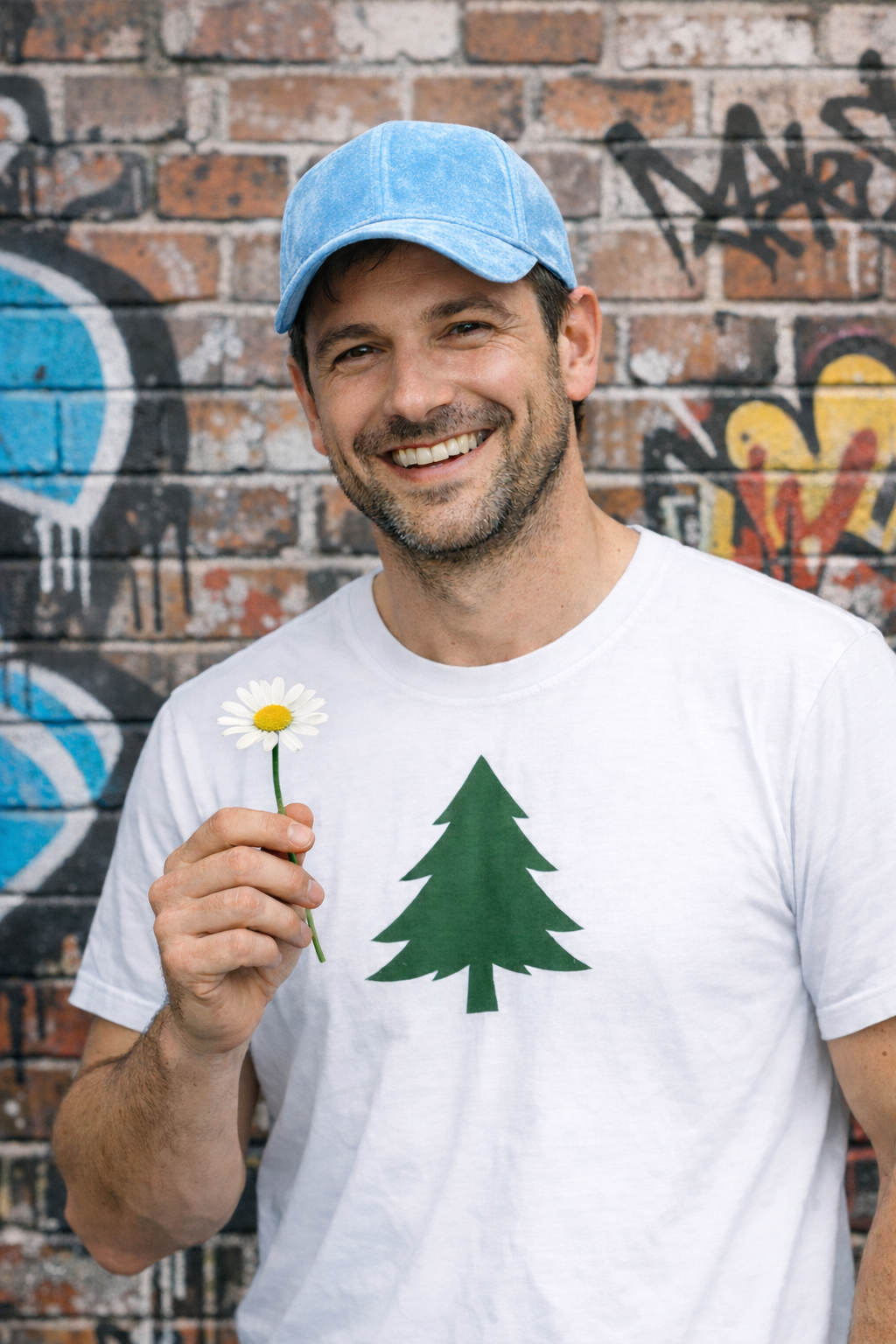 A smiling man in a blue cap and white T-shirt with a green pine tree print, standing against a graffitied brick wall, holding a daisy up beside his face