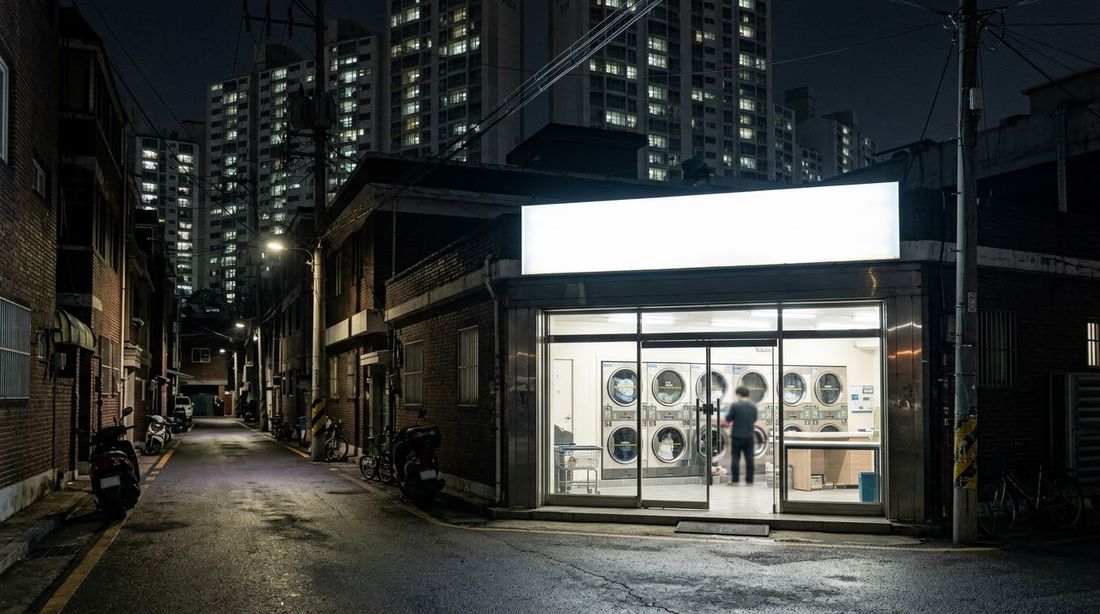 A lit self-service laundry storefront on a quiet Korean residential street at night