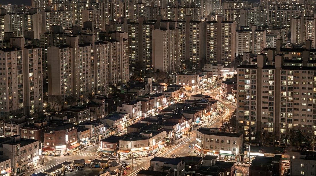 A dense Korean residential district at night, apartment towers rising over lit street-level shops
