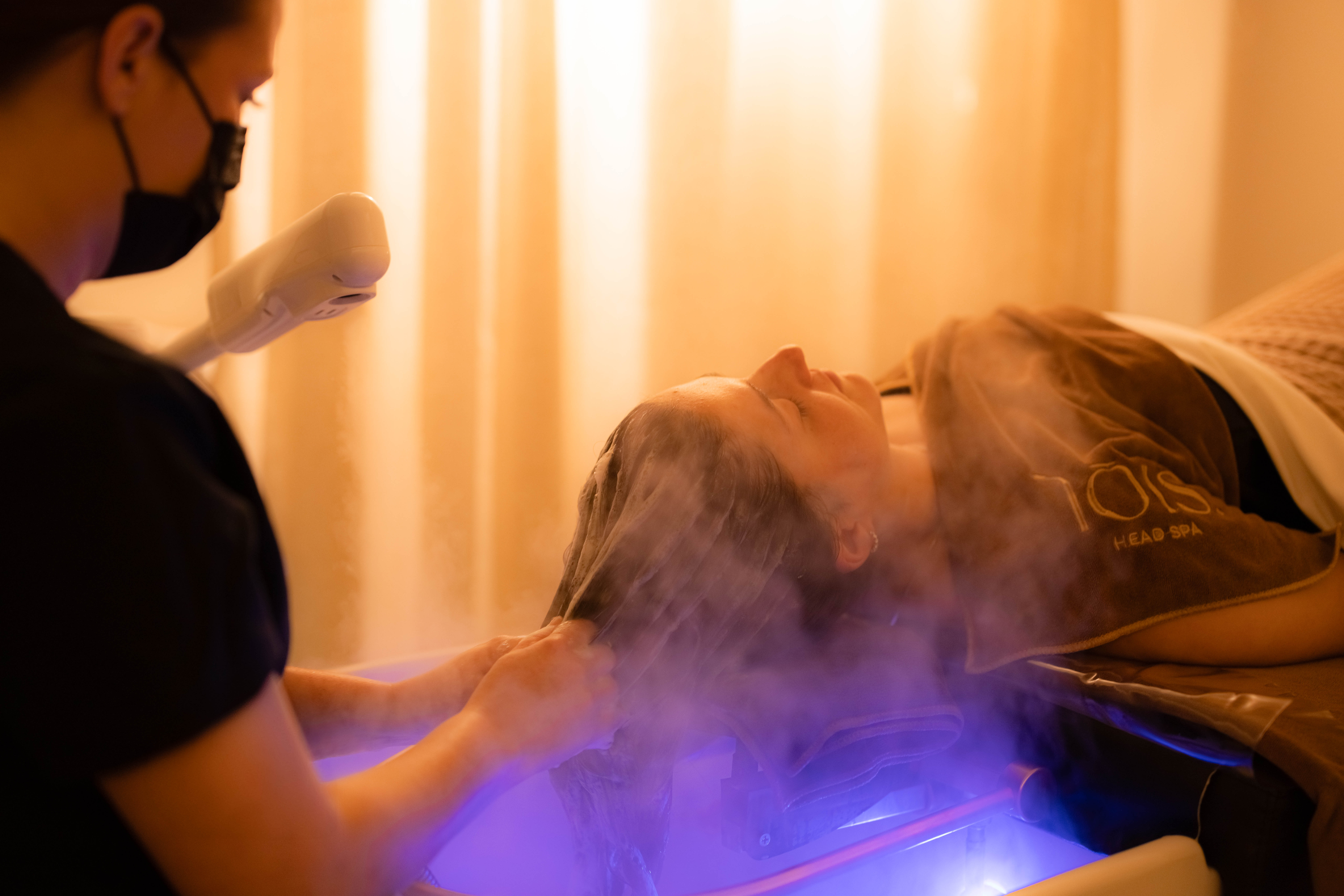Technician holding a steaming herbal ball over a client at the blue-lit basin at Lois Head Spa