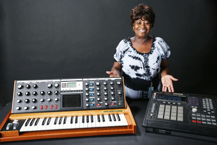 Maureen "Ma Dukes" Yancey smiling with her son J Dilla's MPC 3000