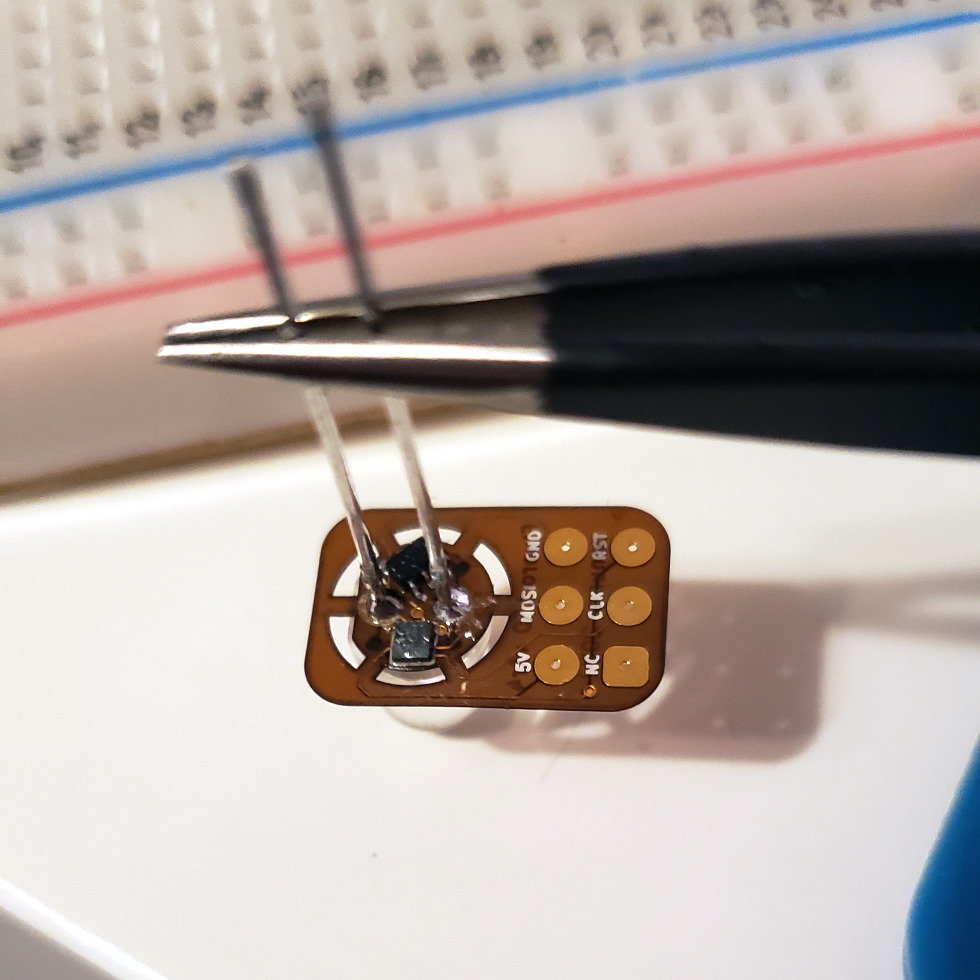 A photograph of a 5mm through-hole LED held in tweezers to expose the small PCB on the underside of the lens, programming pins sticking out