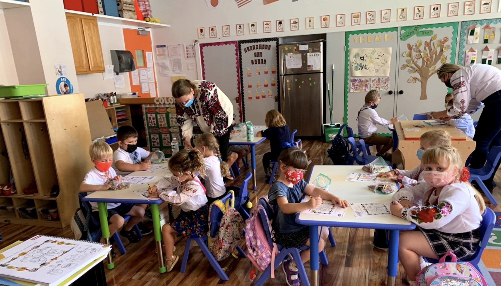 Children dressed in traditional Ukrainian clothes, sitting in a school working on a coloring sheet.