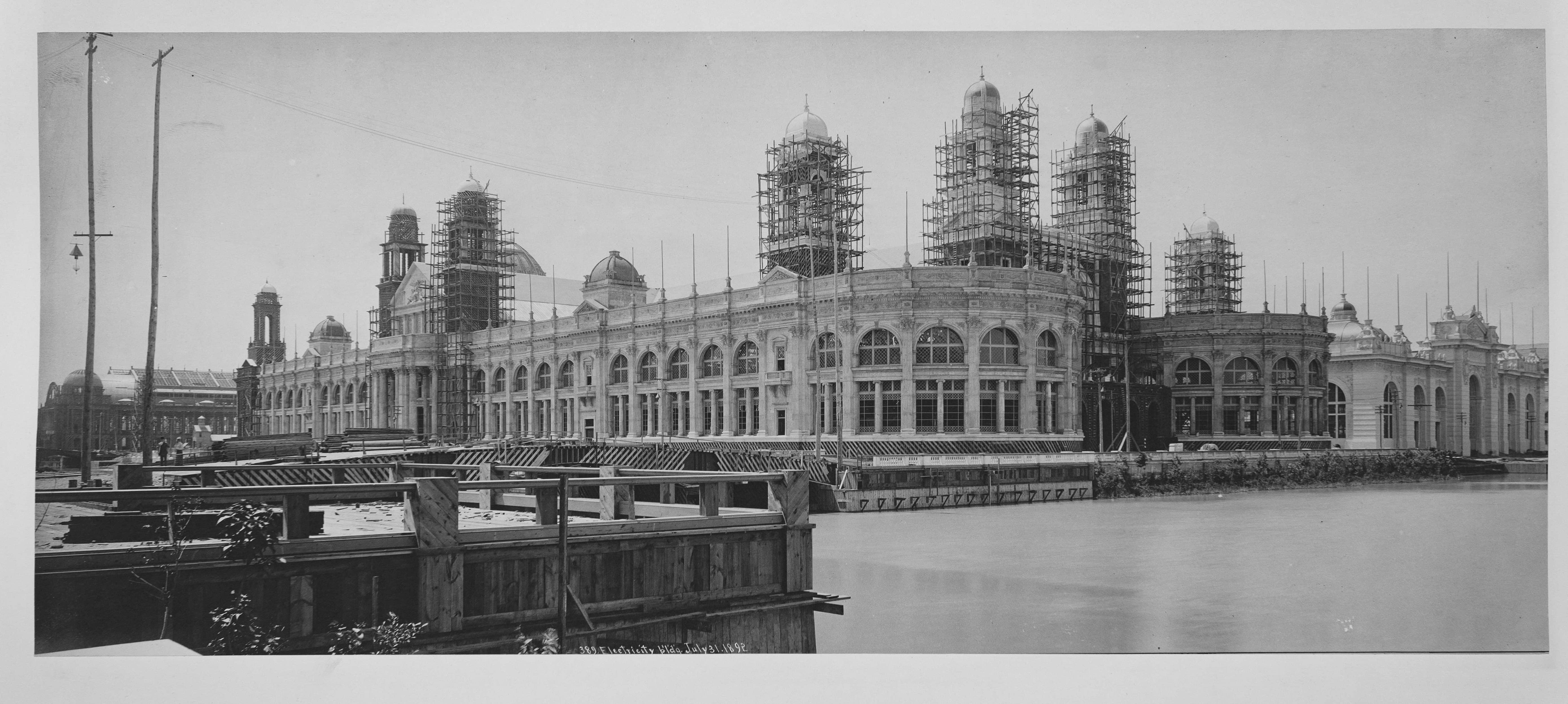 Black and white photograph of a large classical building (Electricity Building) under construction with extensive scaffolding, viewed from across a waterway (1892)