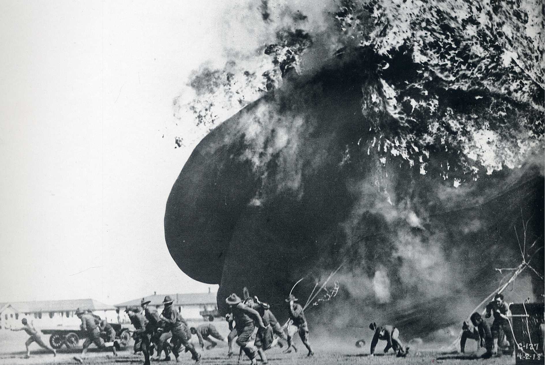 Black and white photograph of military personnel fleeing a large, burning balloon (1918)