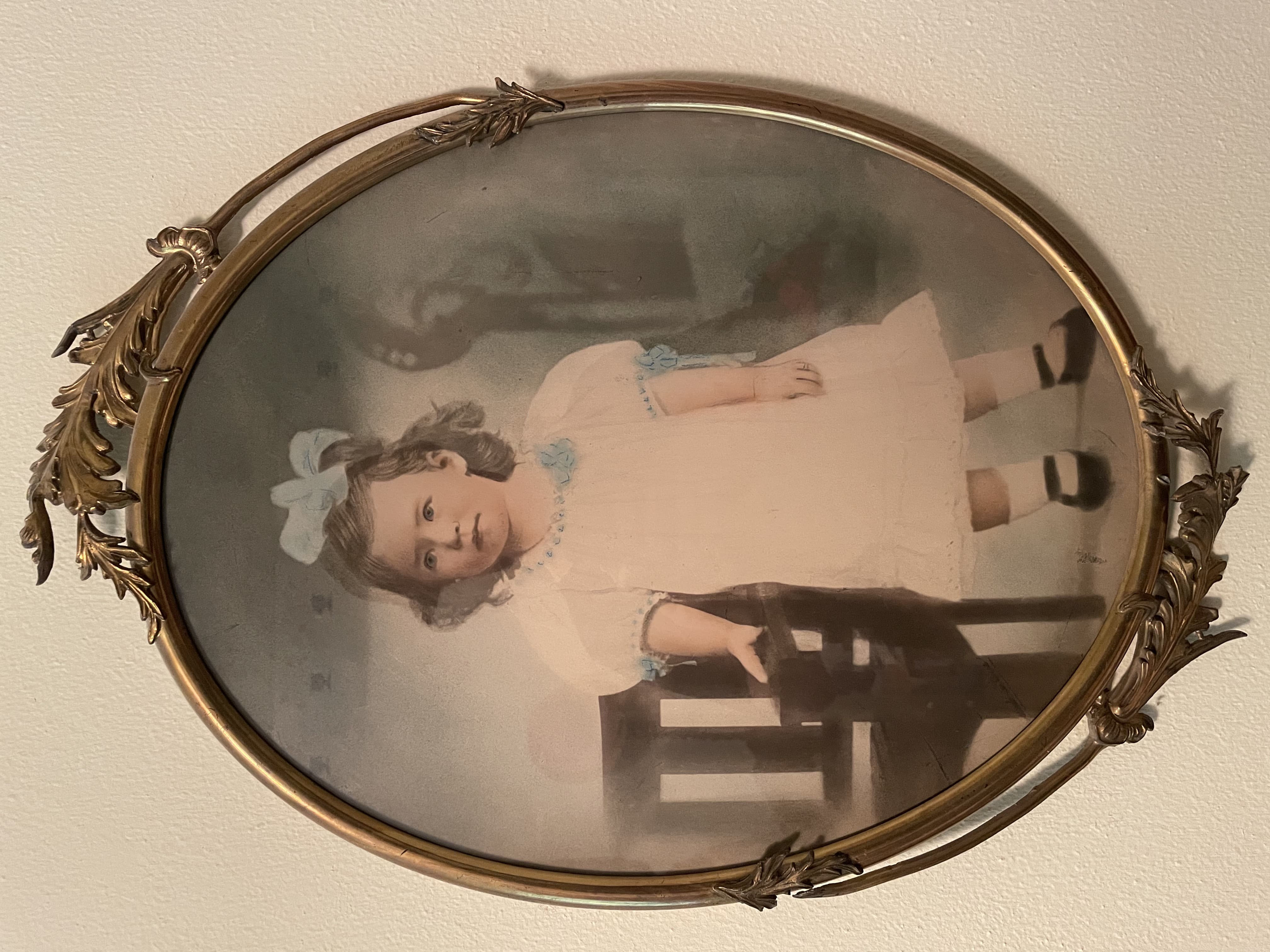 Hand-colored portrait of a young girl in a white dress and blue bow, standing next to a chair