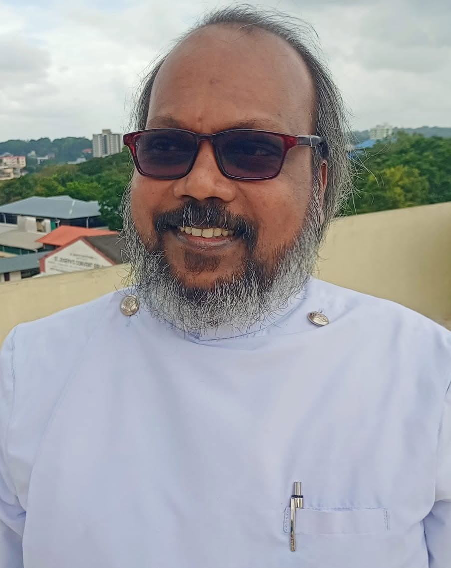 Portrait of Rev. Dr. Y. T. Vinayaraj smiling while wearing glasses and a white clerical robe, standing outdoors with trees, buildings, and a cloudy sky in the background.