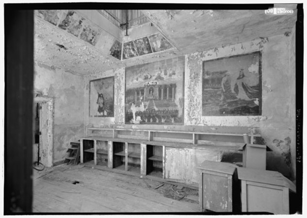 Image of abandoned chaplain's office with religious murals on the wall surrounded by peeling paint and empty cabinets.