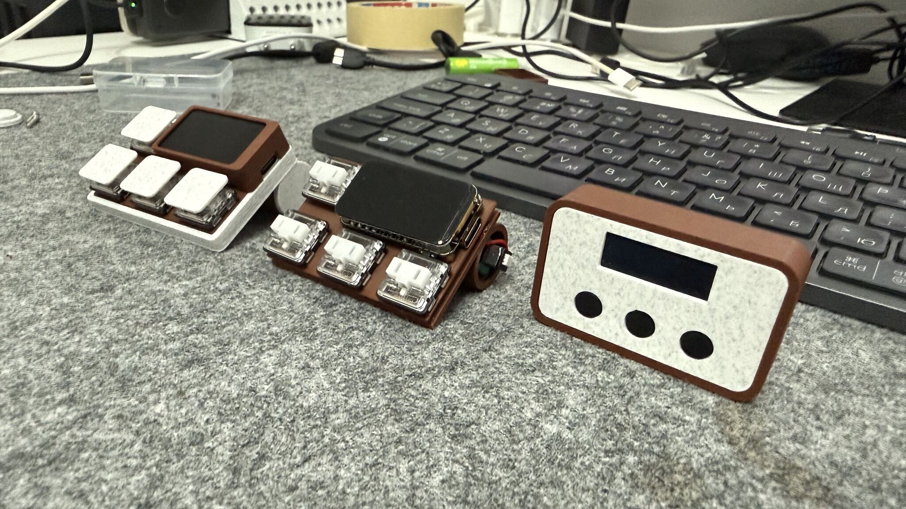 Both Claude Mate builds on a desk — the cordless ESP32-S3 keypad with its colour screen, the same keypad opened up around its cell, and the original Arduino Nano box with its OLED