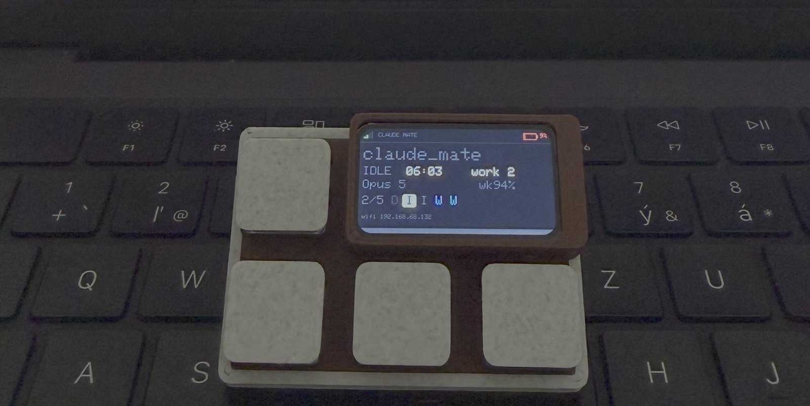 The ESP32-S3 Claude Mate sitting on a laptop keyboard, its colour screen showing a live session
