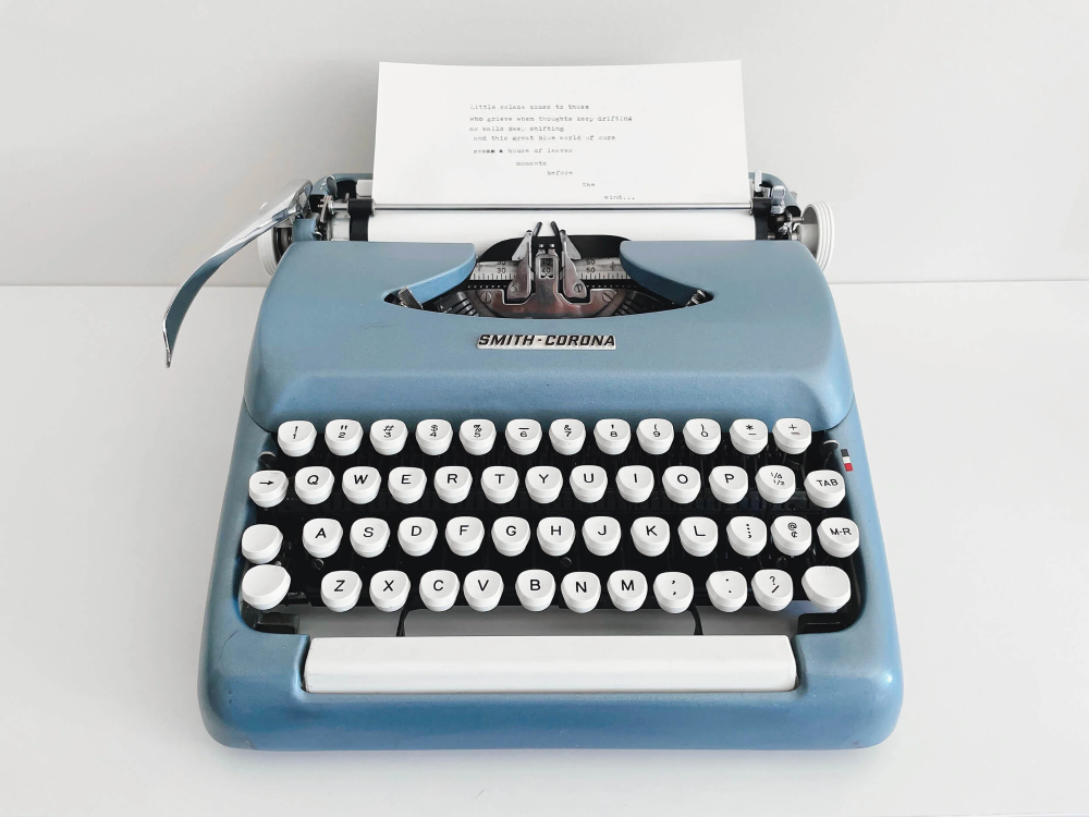 Máquina de escrever vintage azul sobre uma superfície branca, com teclado de teclas redondas brancas e uma folha de papel posicionada no rolo com texto digitado.