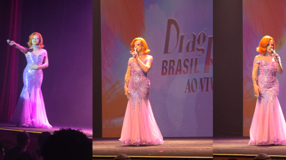 Poseidon Drag Poseidon: ruiva com vestido longo rosa brilhante, estilo glamouroso, segurando microfone e se apresentando de forma mais clássica. iluminação quente e fundo com o telão parcialmente visível.