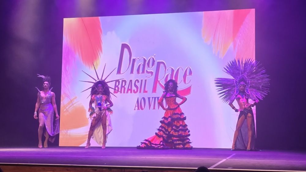 Quatro performers em um palco, alinhados lado a lado, usando figurinos elaborados com penas, estruturas grandes e cores vibrantes. ao fundo, um telão com “Drag Race Brasil ao vivo”. iluminação em tons de roxo e rosa.