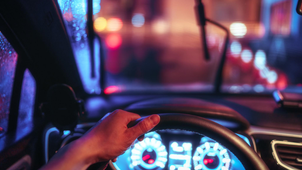 Visão do motorista dentro de um carro à noite, com uma mão no volante; para-brisa com gotas de chuva e luzes desfocadas da cidade ao fundo.
