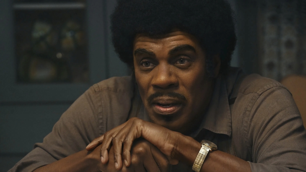 Close no rosto de um homem sentado à mesa, apoiando o queixo sobre as mãos entrelaçadas. Ele tem cabelo volumoso em estilo afro, bigode e expressão carregada, com olhar fixo e levemente cansado, como se estivesse refletindo ou processando algo difícil. A iluminação é quente e suave, vindo lateralmente, criando sombras que reforçam as marcas do rosto e a profundidade da cena. Ele usa uma camisa casual de tom neutro e um relógio metálico no pulso. O fundo está desfocado, sugerindo um ambiente doméstico, possivelmente uma cozinha ou sala de jantar, com elementos como armários e objetos ao fundo. A composição é íntima, focada na expressão emocional do personagem.