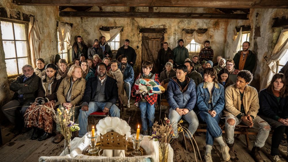 Cena da série From mostrando dezenas de moradores reunidos em um velório dentro de uma casa antiga e desgastada. O ambiente é escuro e melancólico. No centro da imagem, um garoto segura um cobertor colorido enquanto os demais personagens observam em silêncio, transmitindo luto, exaustão e tensão coletiva.