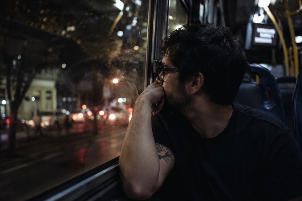 Homem sentado sozinho em um ônibus durante a noite, apoiando o rosto sobre a mão enquanto observa a cidade pela janela. A iluminação está baixa do interior do veículo, luzes desfocadas da rua ao fundo. O reflexo do rosto aparece parcialmente no vidro. Ele usa camiseta preta e óculos, com uma tatuagem visível no braço.