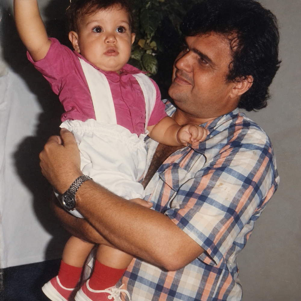Homem segura uma criança pequena no colo e olha para ela sorrindo, enquanto a criança, vestida de rosa e branco, mantém um dos braços levantado.