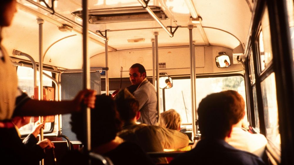 Interior de um ônibus antigo, com passageiros sentados e em pé, barras metálicas de apoio e iluminação alaranjada entrando pelas janelas.